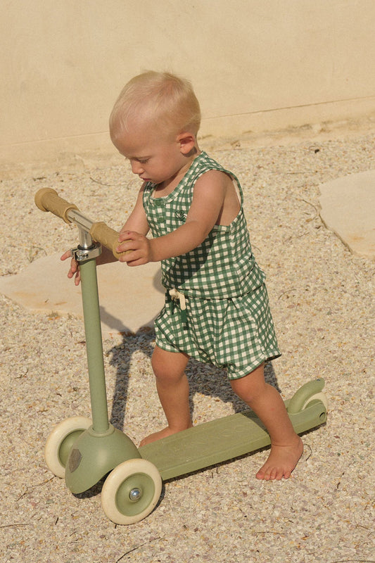 Child in a green checkered outfit playing with a green scooter on a sandy surface.